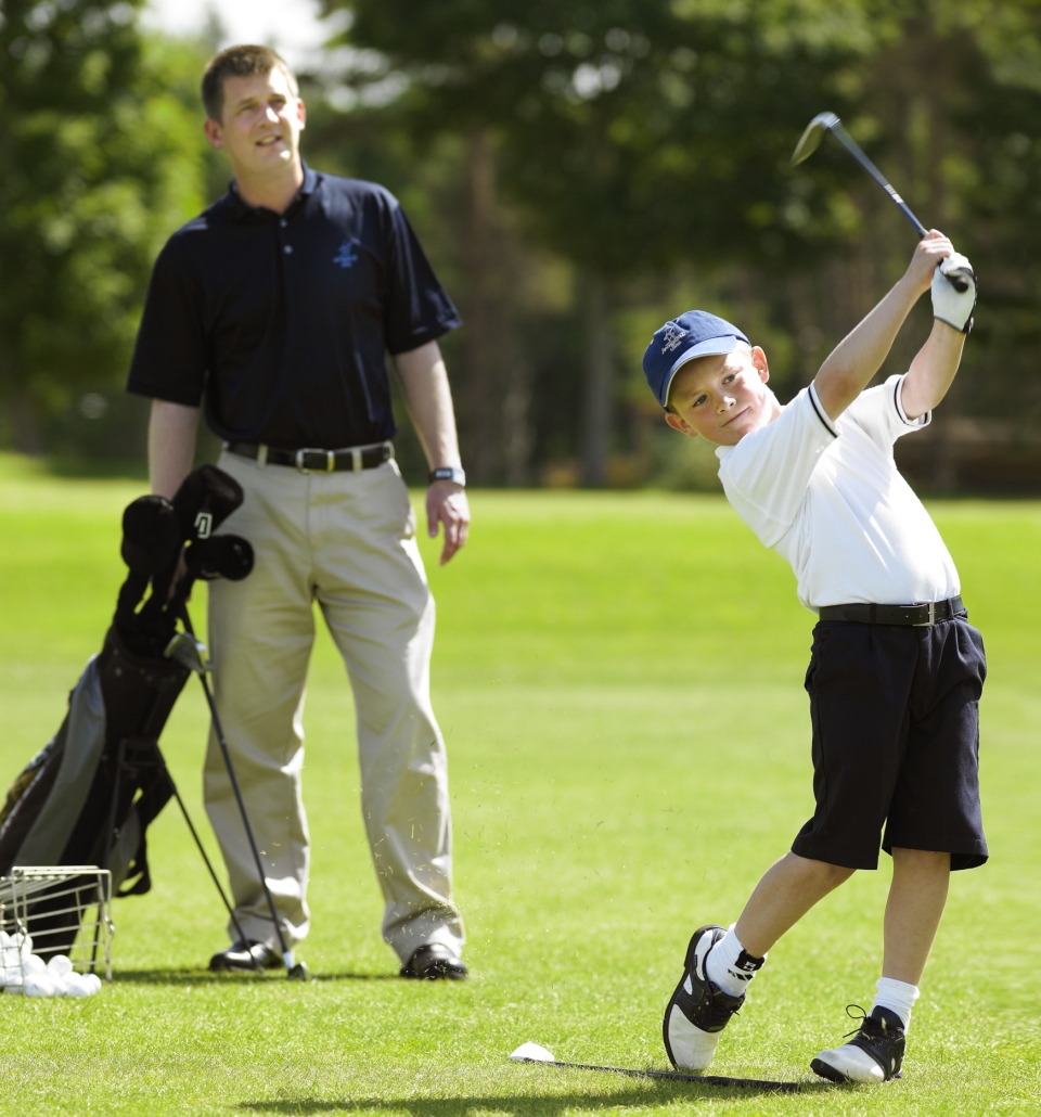 a kid swinging his golf club with his dad looking at him from behind