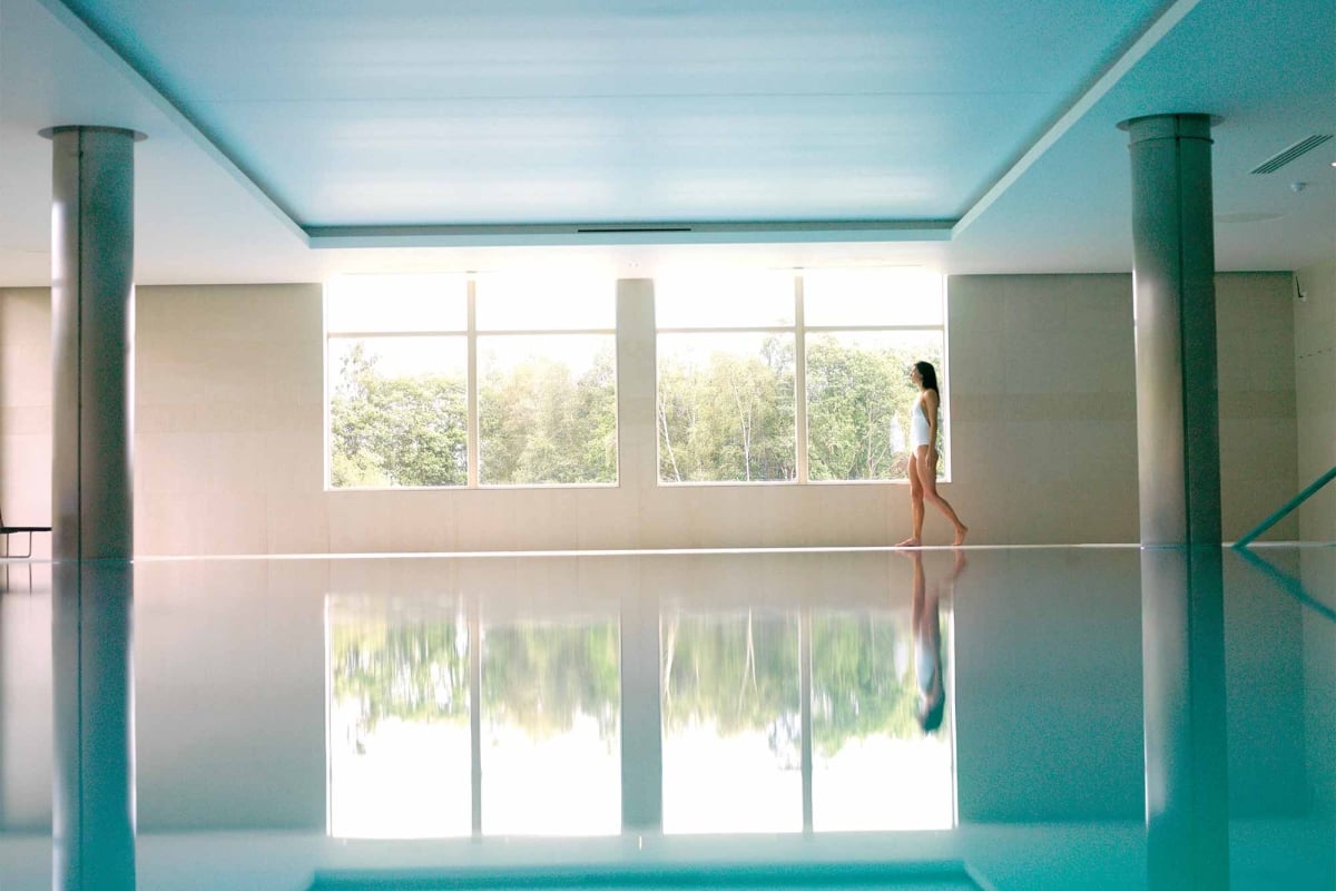 a woman walking along the side of a indoor pool looking outside the window