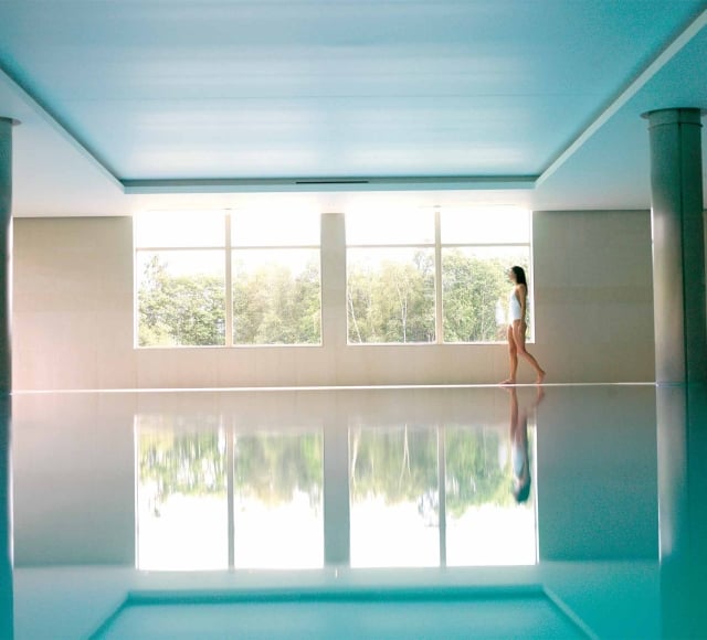 a woman walking along the side of a indoor pool looking outside the window