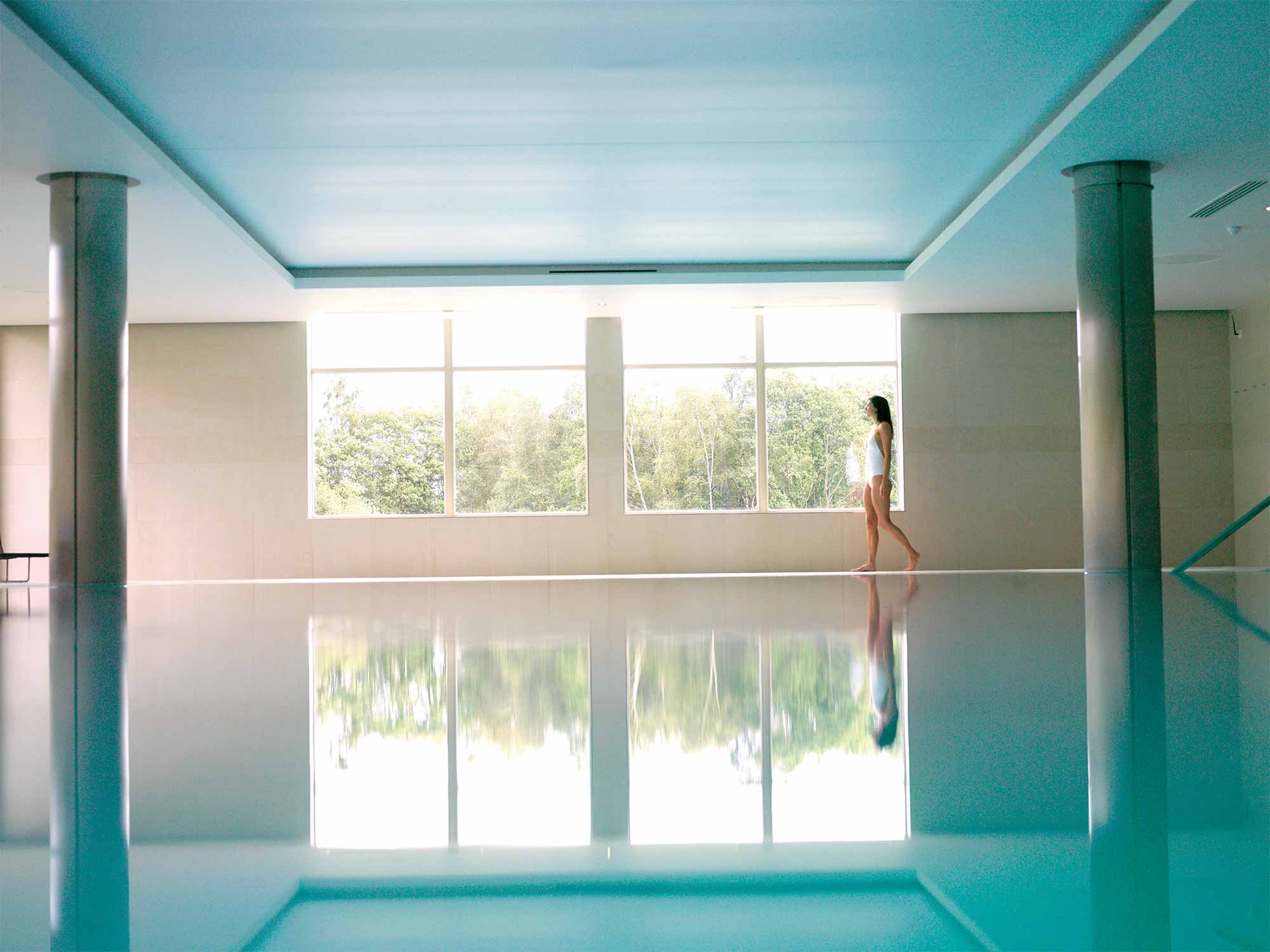 a woman walking along the side of a indoor pool looking outside the window