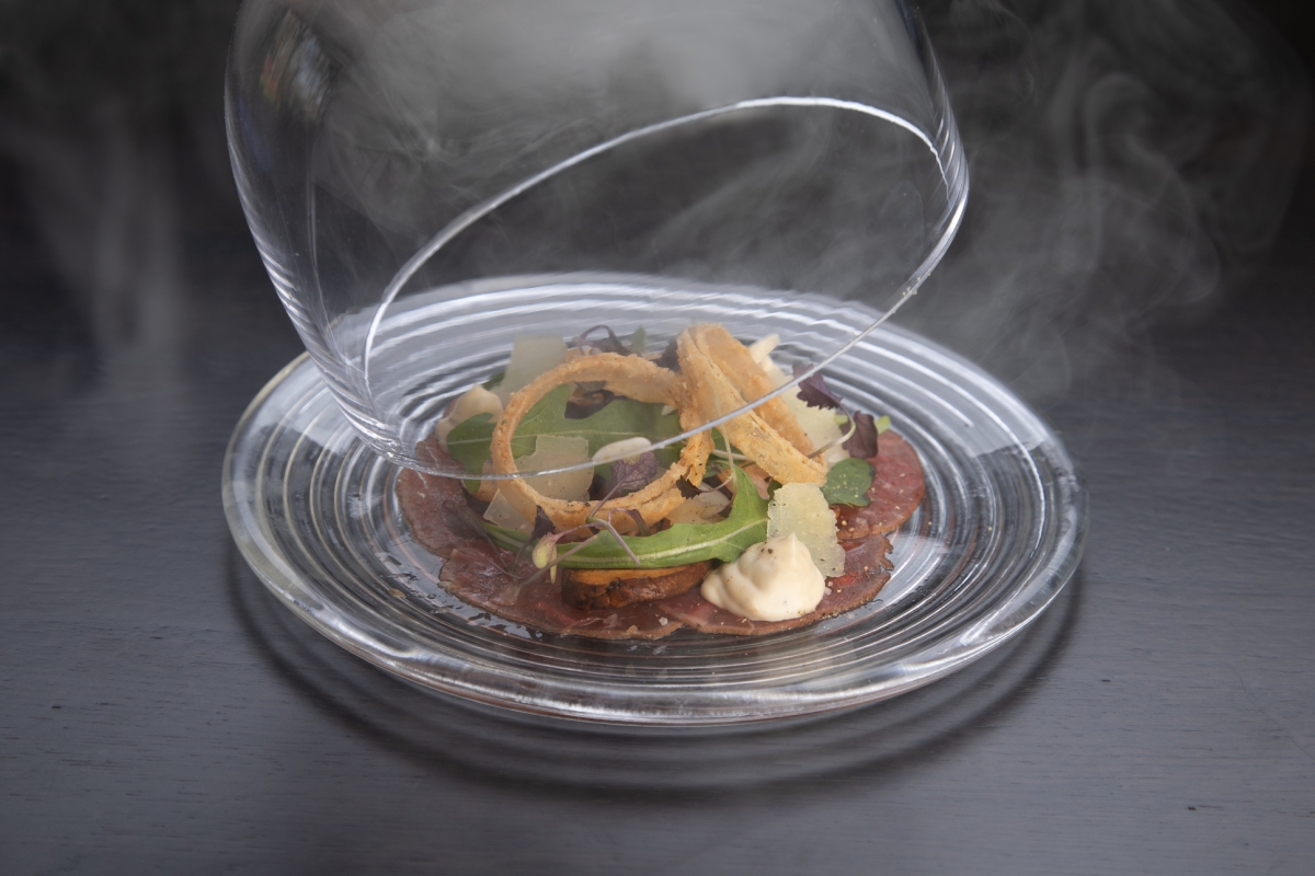 A server lifting a glass covering off of a plate of carpaccio of beef at La Vista restaurant at Cameron House.