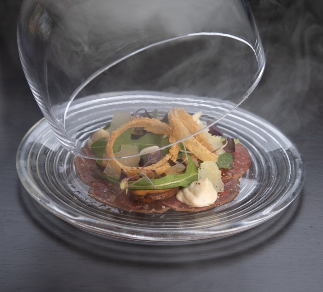 A server lifting a glass covering off of a plate of carpaccio of beef at La Vista restaurant at Cameron House.