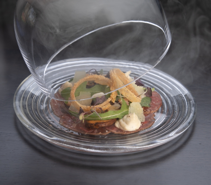 A server lifting a glass covering off of a plate of carpaccio of beef at La Vista restaurant at Cameron House.