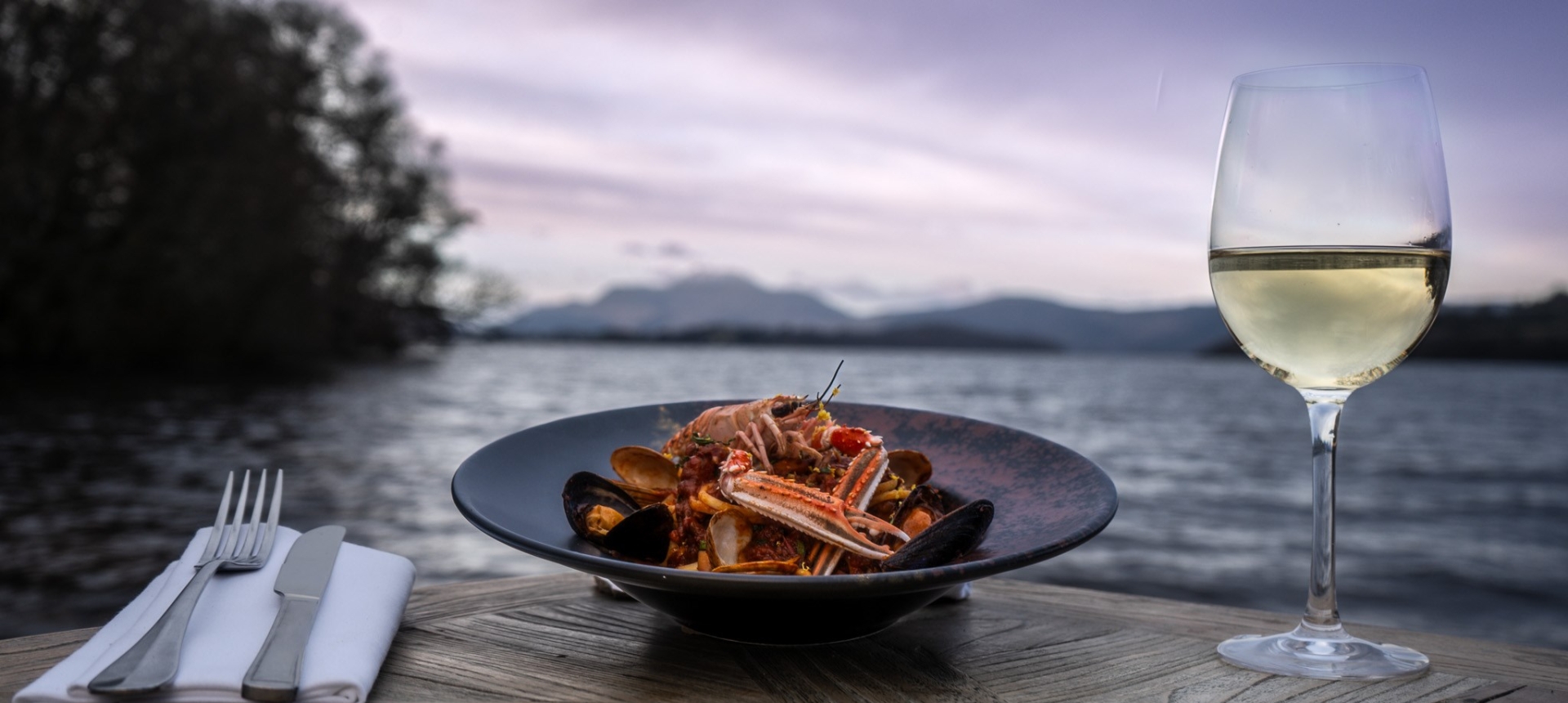 a beautiful seafood dish sitting next to silverware and champagne out doors in front of the water