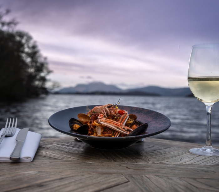 a beautiful seafood dish sitting next to silverware and champagne out doors in front of the water