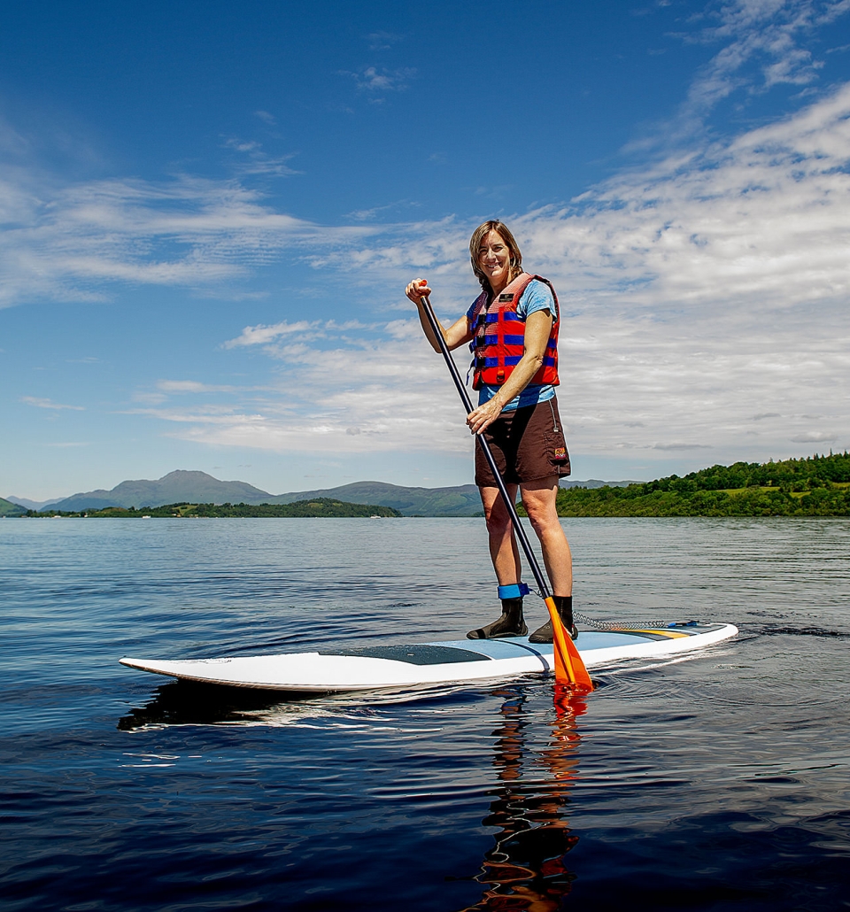 a woman standing on a paddle board in the middle of the water with bright green trees and hills behind her