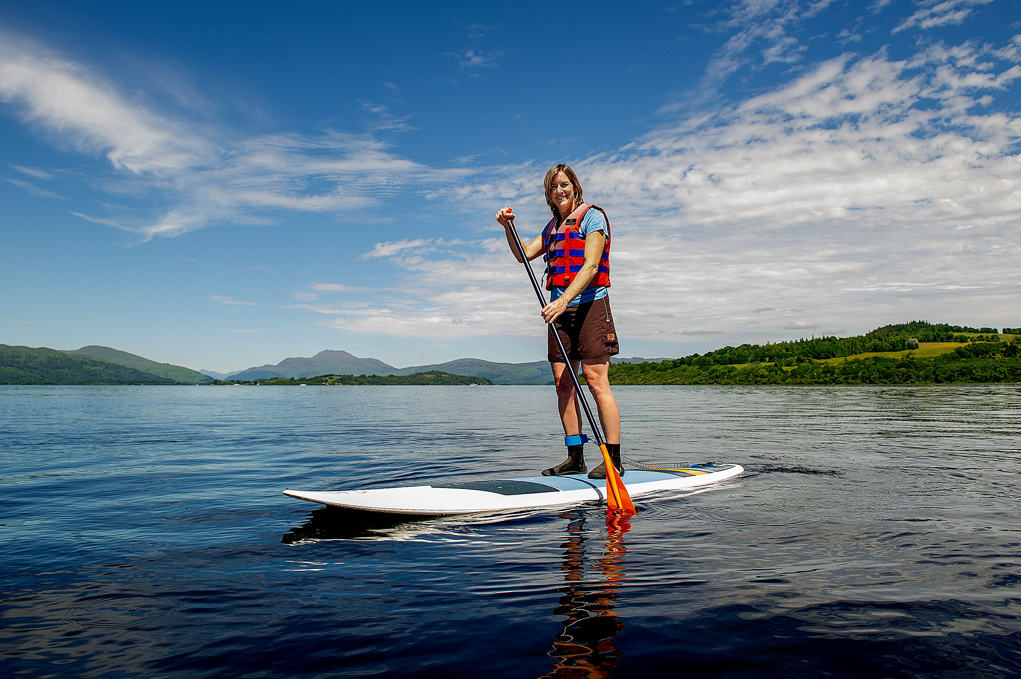 a woman standing on a paddle board in the middle of the water with bright green trees and hills behind her