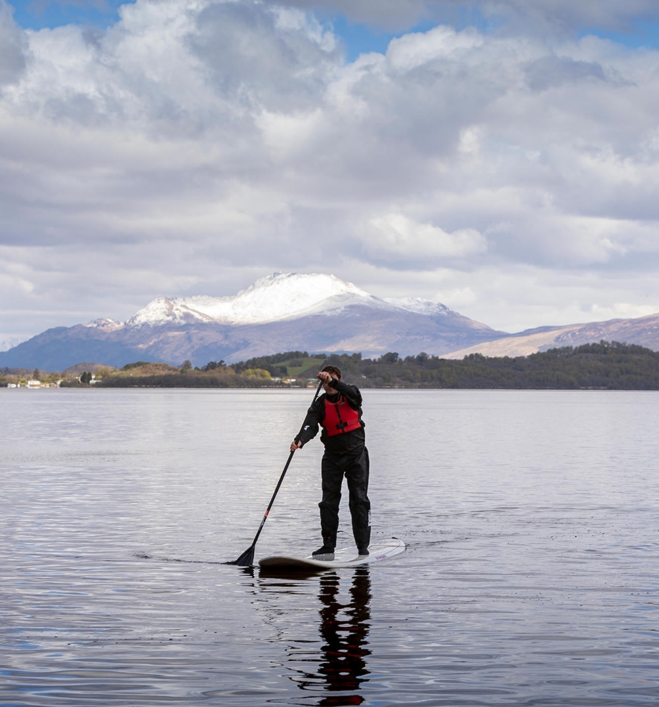 a man standing on a paddle board paddling across the water with snowy mountains in the background