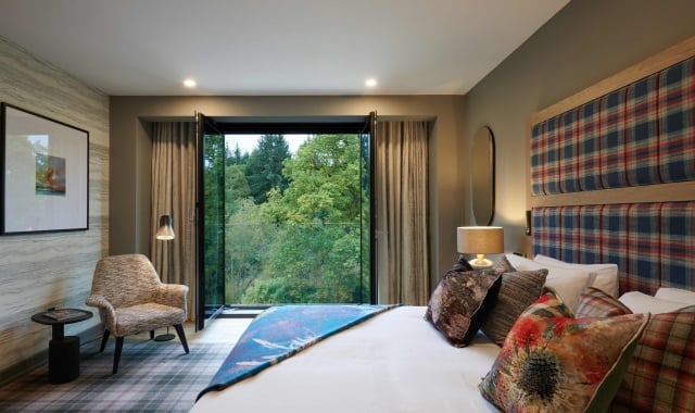 Hotel room with bed, chair, and view of lush green trees from window.