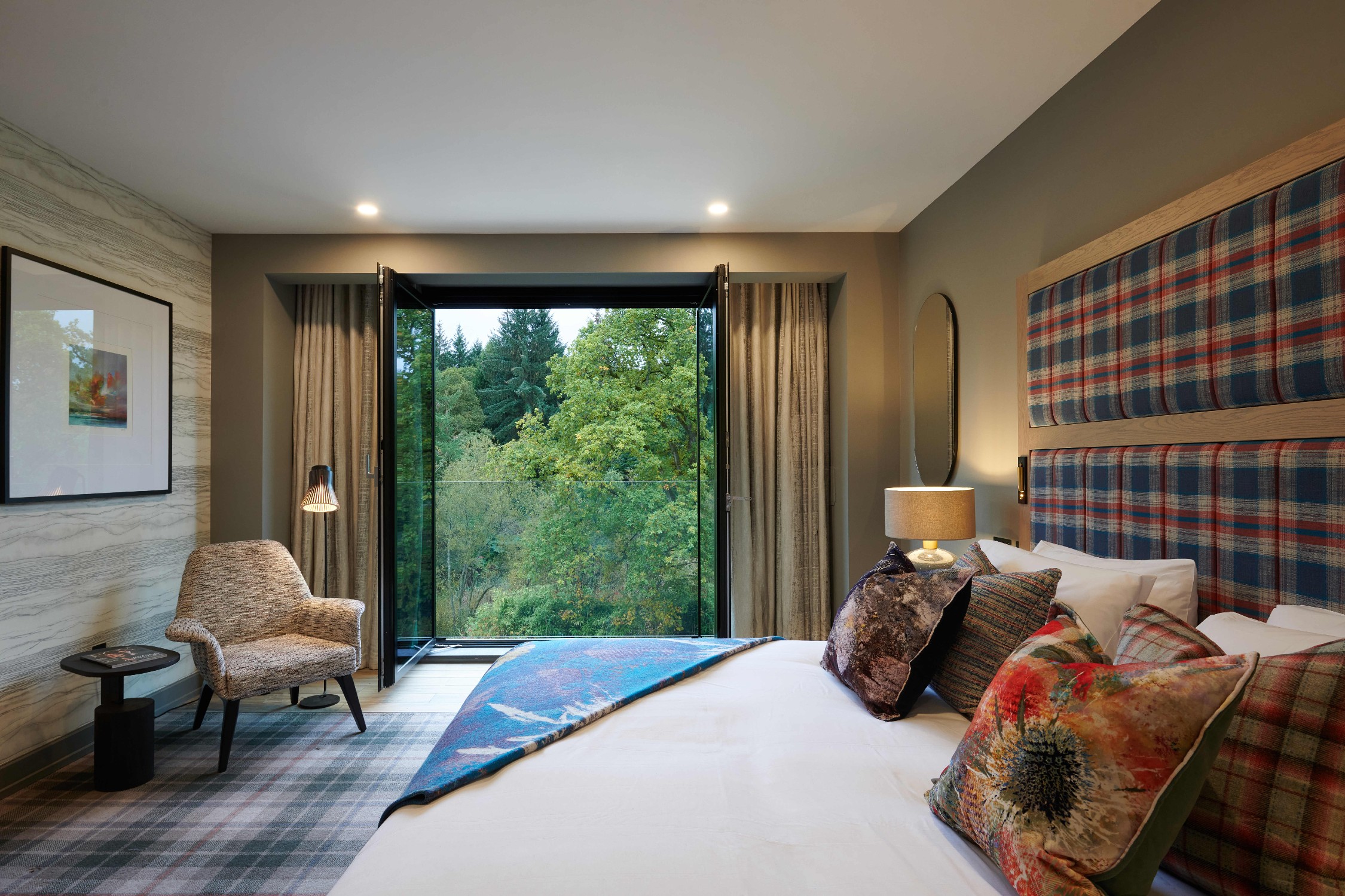 Hotel room with bed, chair, and view of lush green trees from window.